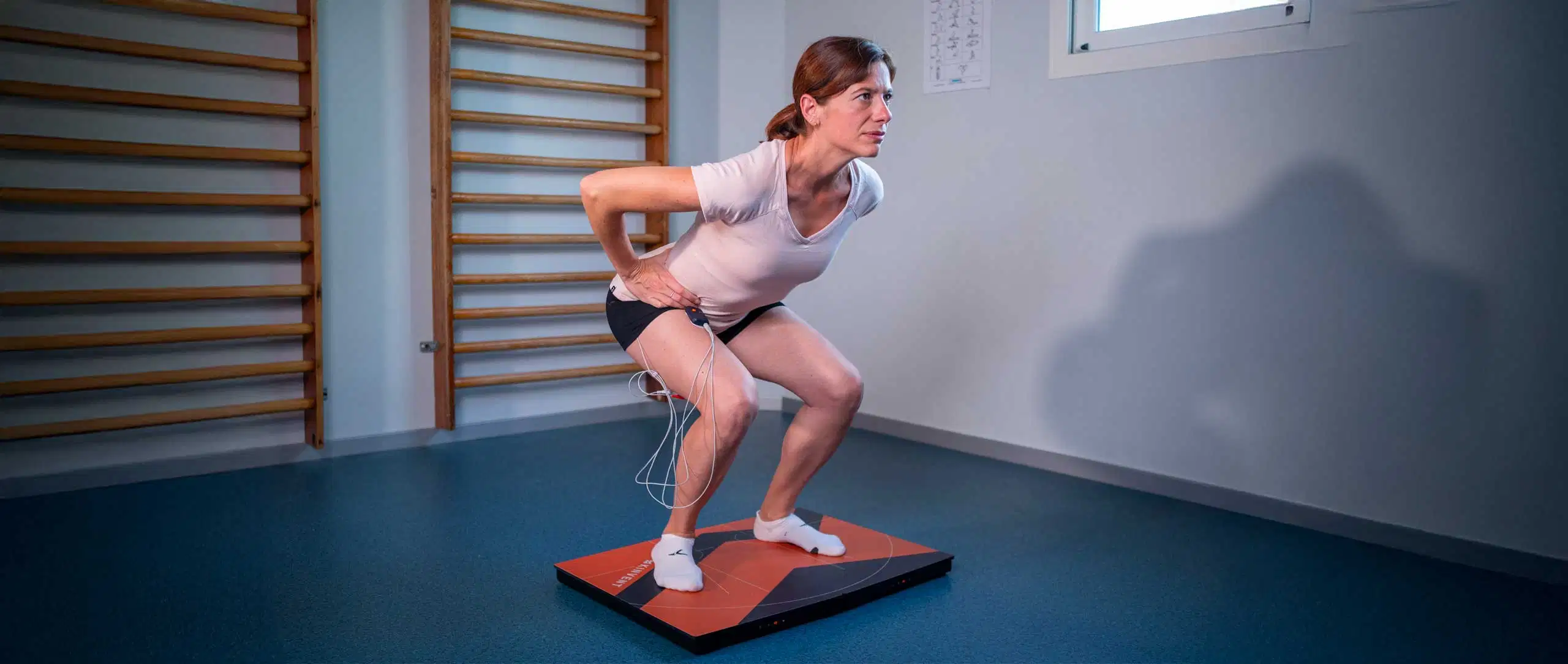 A woman performing a squat test on a pair of K-Deltas force plates while wearing K-Myo electromyography (EMG) sensors with wires on her leg to measure muscle activity