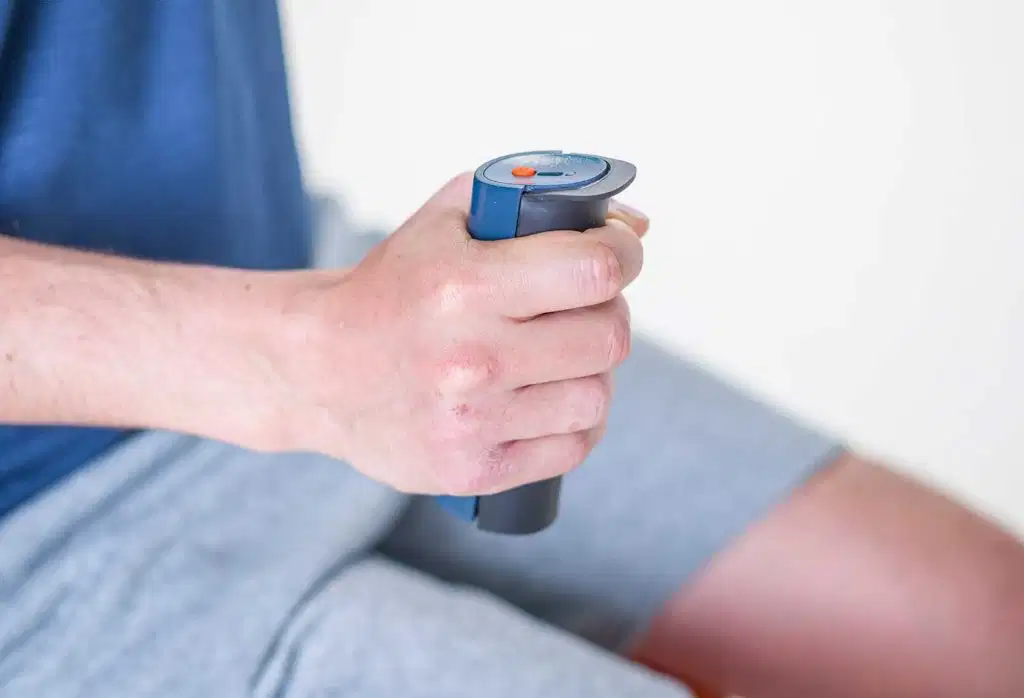 A side view of a man's arm and hand squeezing a blue and grey K-Grip dynamometer to measure grip strength