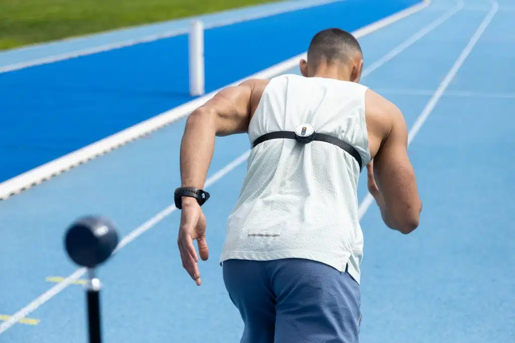 Rear view of a runner starting a sprint on a blue running track, wearing a Kinvent sensor strap across his upper back to analyze movement dynamics