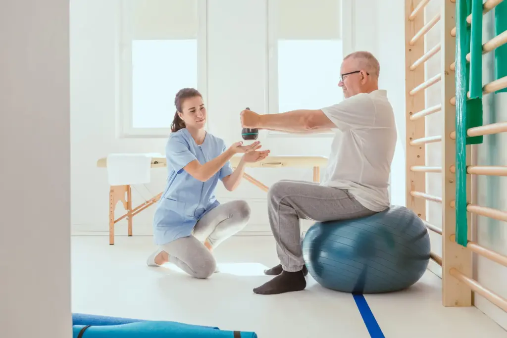Un patient senior s'entraîne sur un ballon de gym bleu lors d'une séance de rééducation avec une kinésithérapeute en cabinet