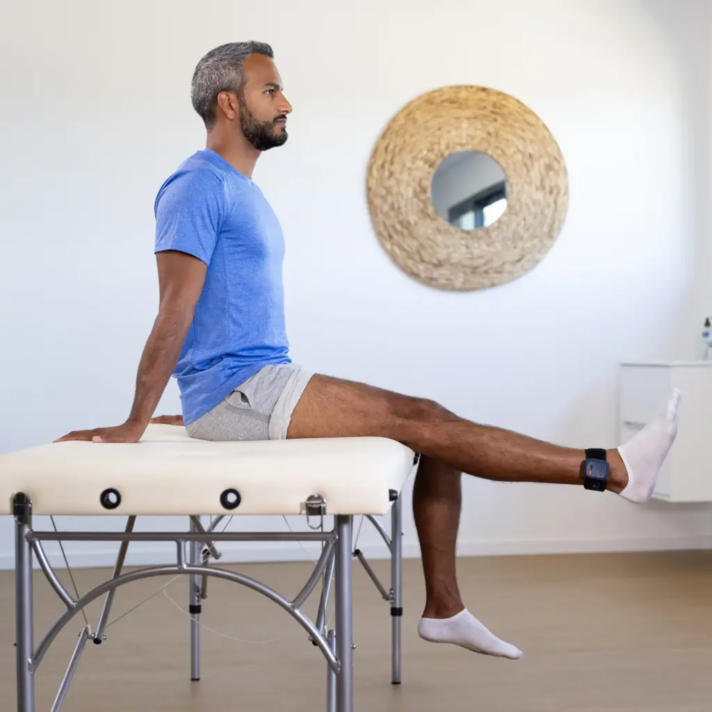 Un patient assis sur une table d'examen effectuant un exercice d'extension de la jambe avec un capteur connecté Kinvent à la cheville pour la rééducation du genou