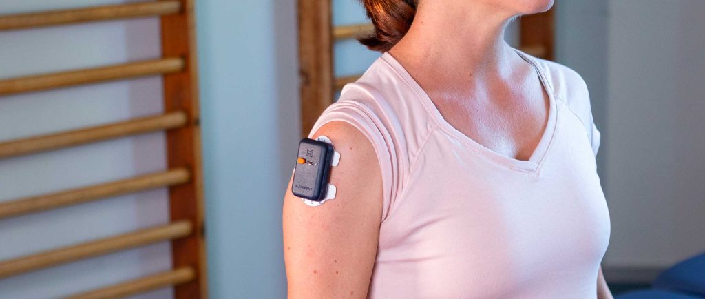 A close-up profile shot of a woman with a Kinvent K-Myo wireless EMG sensor attached to her shoulder muscle to track muscle activation during a physical therapy session