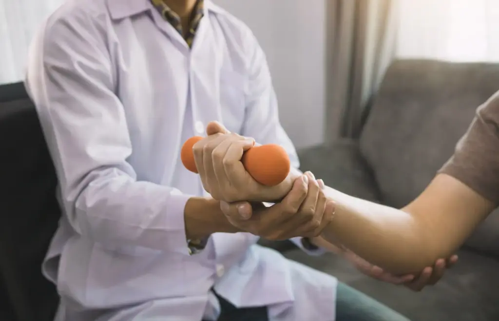 Gros plan sur un kinésithérapeute en blouse blanche soutenant le bras d'un patient qui serre un petit haltère orange, illustrant le travail de la force de préhension et la rééducation de la main