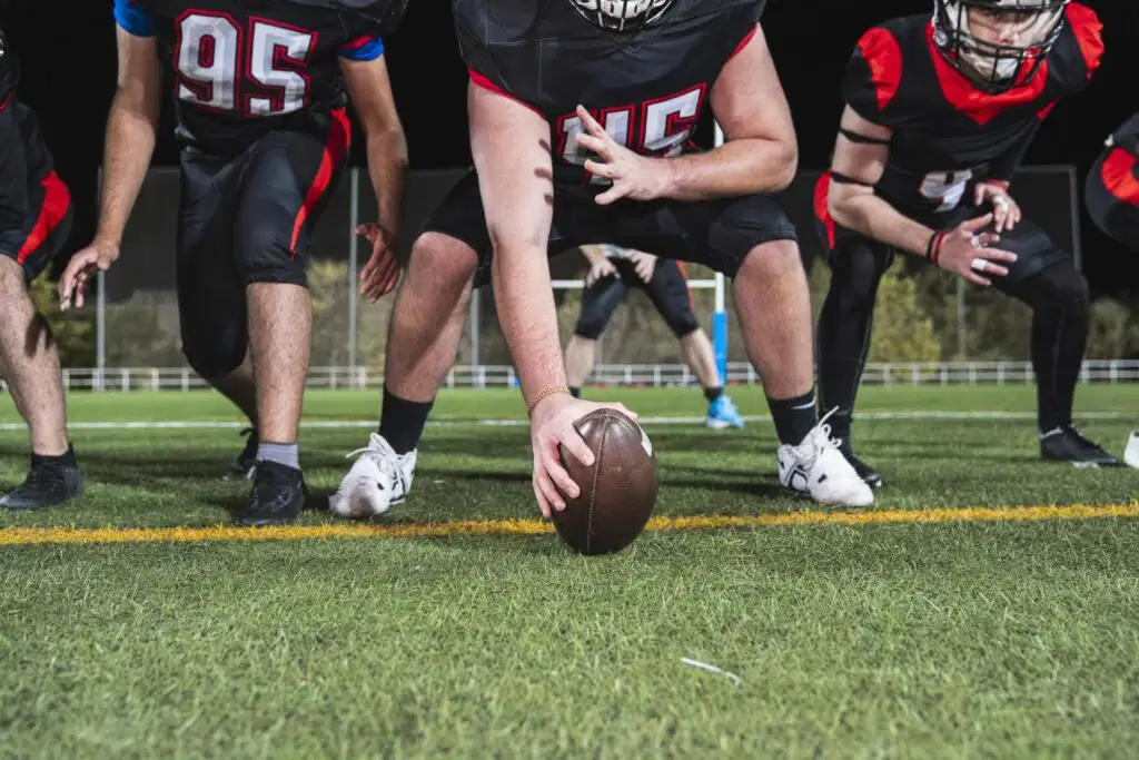 American football players in black and red uniforms lined up on the field, with the center's hand on the football ready to snap, illustrating sports neck training and concussion prevention context