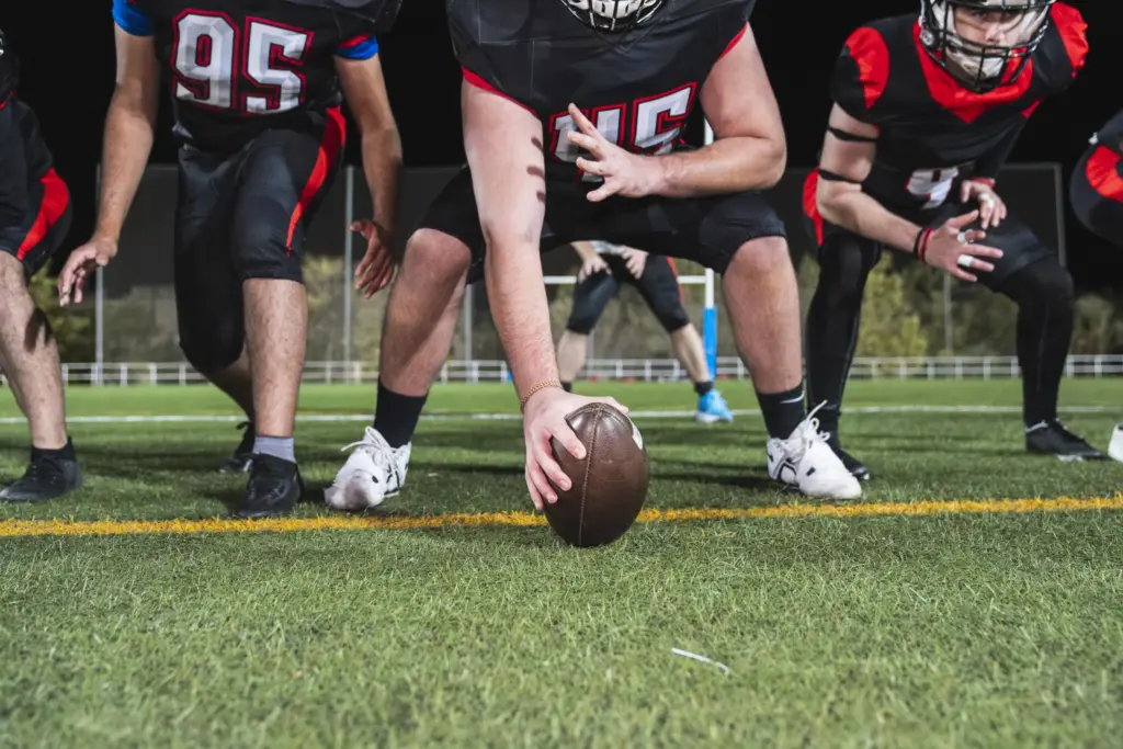 Neck Training for Concussion Prevention: Why Strengthening the Neck Matters in Sports 43 American football players in black and red uniforms lined up on the field, with the center's hand on the football ready to snap, illustrating sports neck training and concussion prevention context