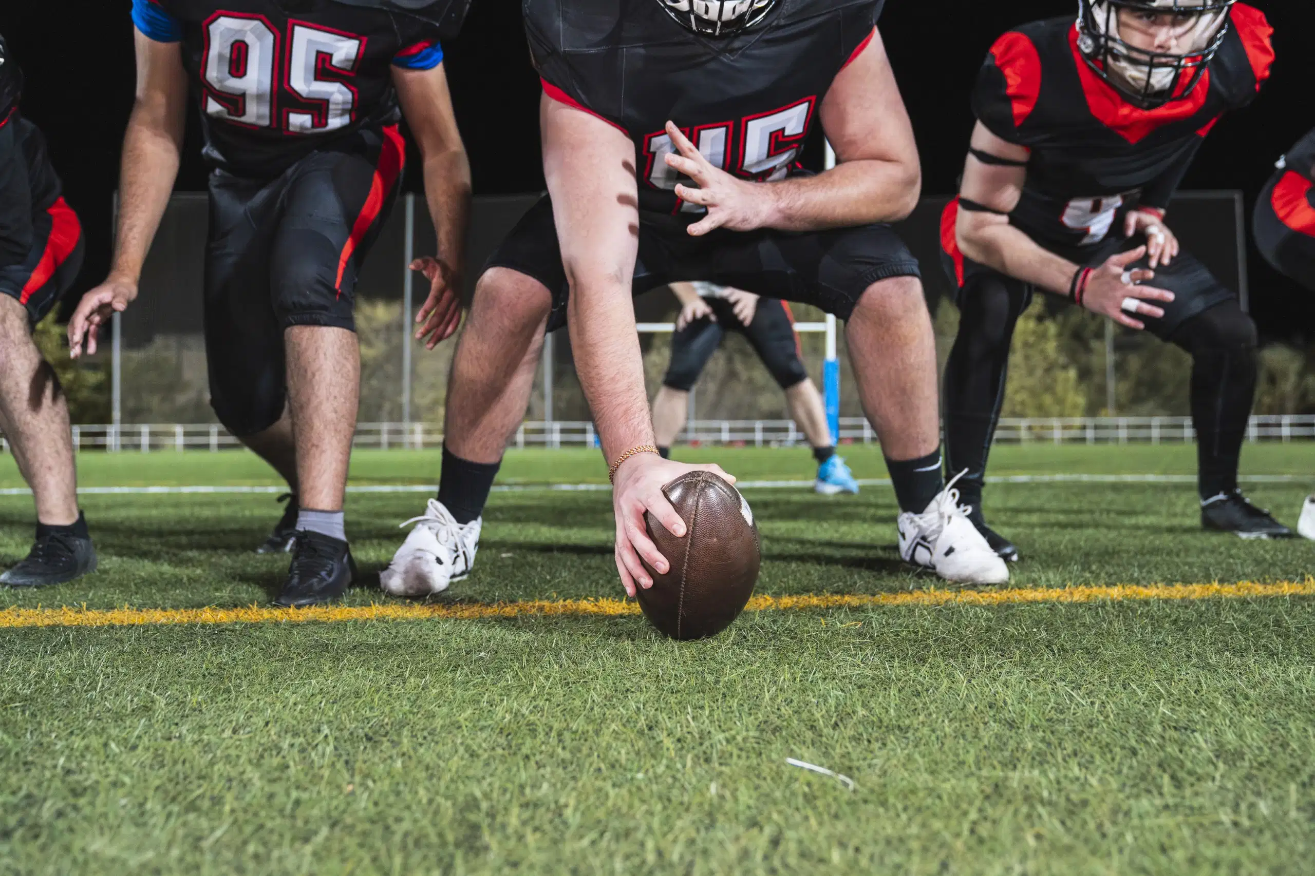 American football players in black and red uniforms lined up on the field, with the center's hand on the football ready to snap, illustrating sports neck training and concussion prevention context