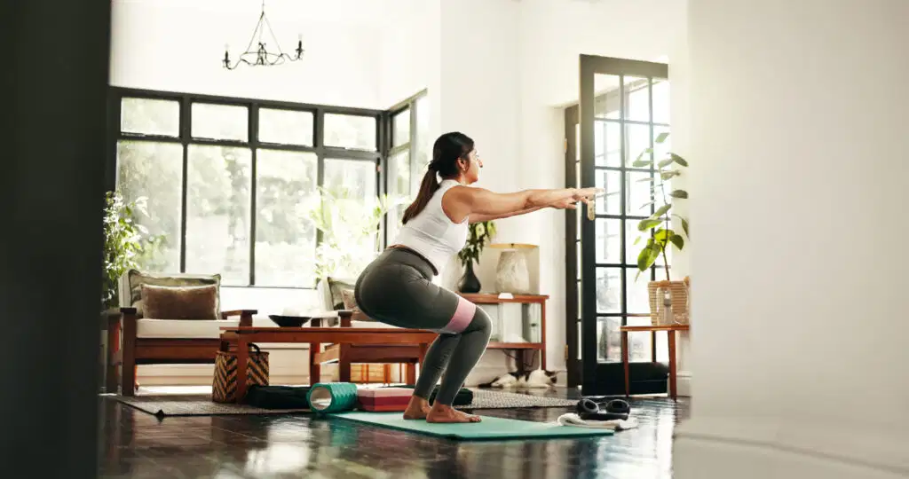 Los cuatro pilares del movimiento humano: movilidad, estabilidad, fuerza y movimiento funcional explicados 1 Woman performing a squat with a resistance band in a living room, demonstrating a lower body strength exercise