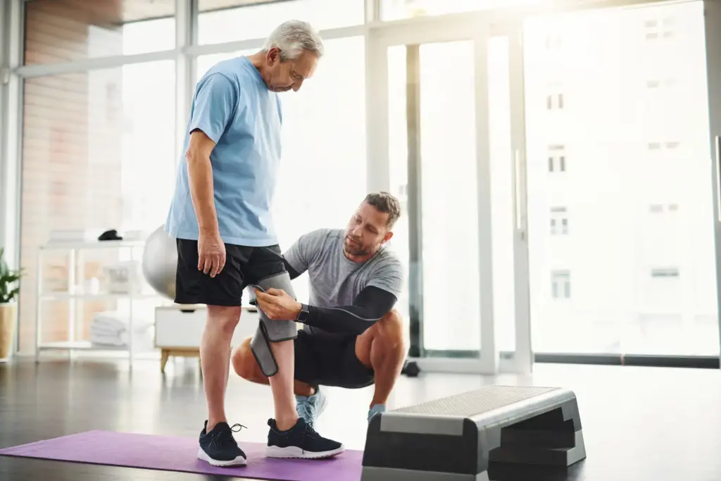 Balanceo postural tras el reemplazo de rodilla: la importancia de la complejidad del equilibrio para prevenir caídas 10 Physical therapist adjusting a knee brace on a senior patient during a rehabilitation session for postural sway and balance after knee replacement