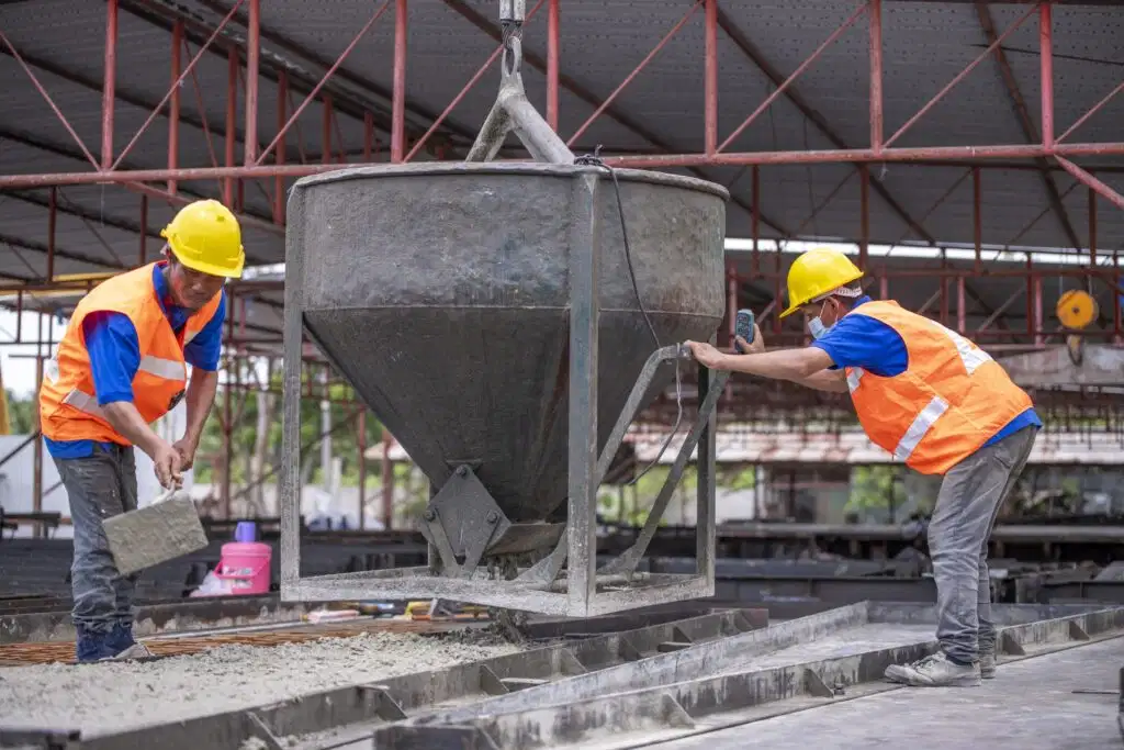 Two construction workers in safety gear bending and lifting to manage a concrete bucket on a site, illustrating functional movement