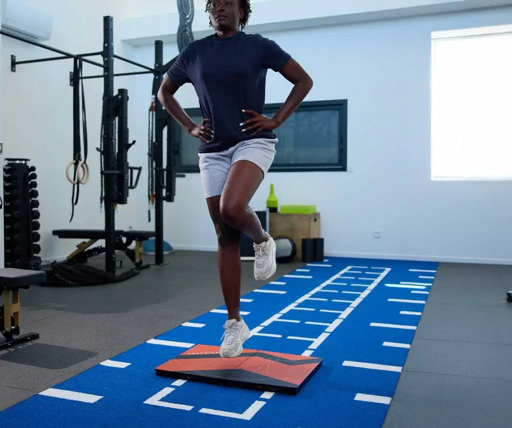 An athlete with hands on her hips performing a single-leg countermovement jump (CMJ) assessment on orange K-Deltas force plates inside a training facility