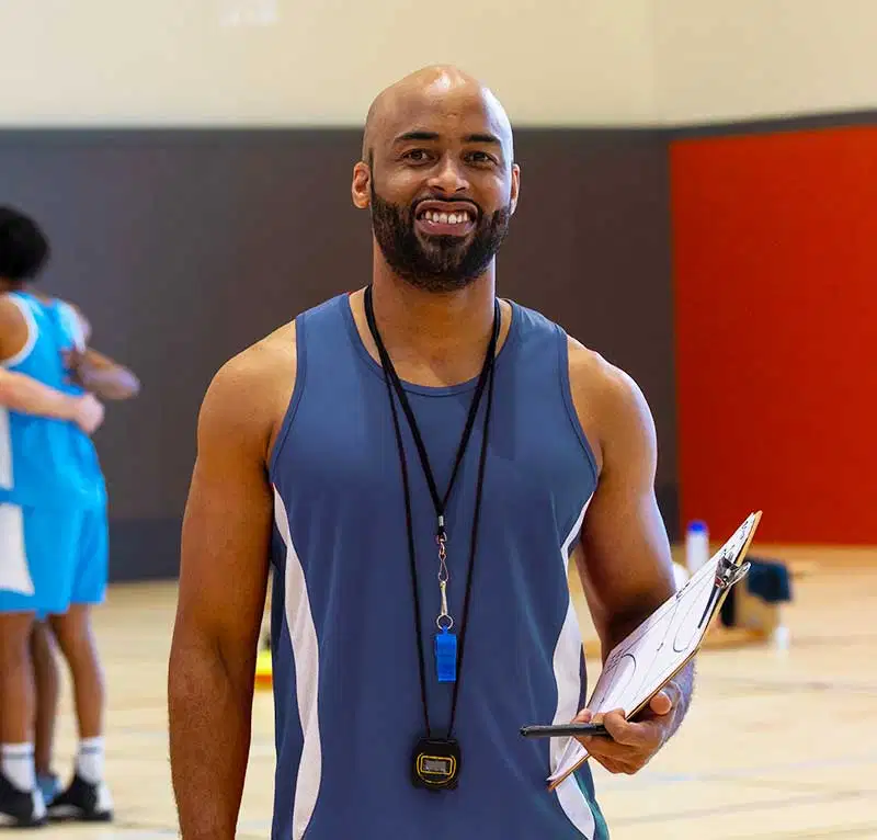 A portrait of a smiling male coach holding a tactical basketball clipboard, wearing a blue tank top, a whistle, and a stopwatch around his neck inside a gym