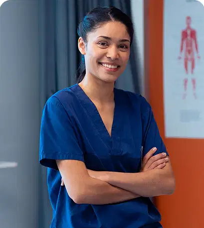 A professional portrait of a smiling female physical therapist or healthcare professional with folded arms, wearing dark blue scrubs inside a clinic room with an anatomical chart on the wall