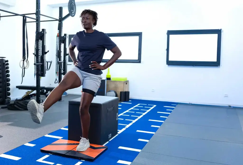 Una atleta con las manos en las caderas realizando una evaluación de sentadilla unipodal sobre un par de plataformas de fuerza K-Deltas naranjas en un centro de entrenamiento