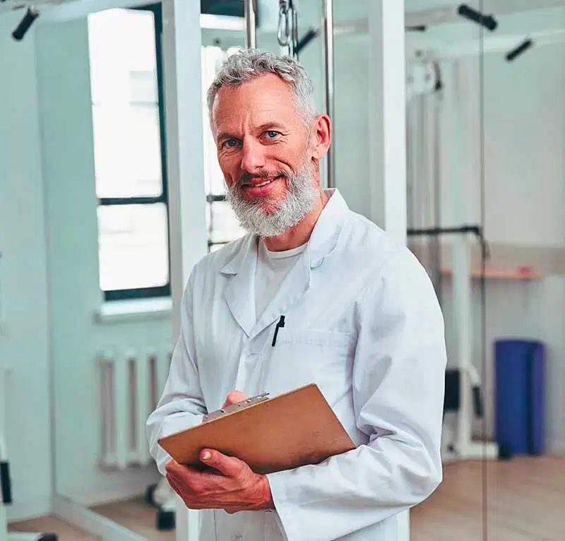 Retrato de un médico mayor sonriente con barba canosa, vistiendo una bata blanca y sosteniendo un portapapeles de madera en una clínica médica o de rehabilitación luminosa.