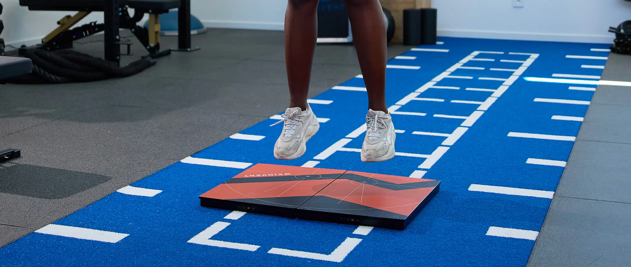 A close-up action shot of an athlete's legs mid-air performing a Countermovement Jump (CMJ) over two large Kinvent K-Deltas force plates positioned on a blue turf track inside a gym
