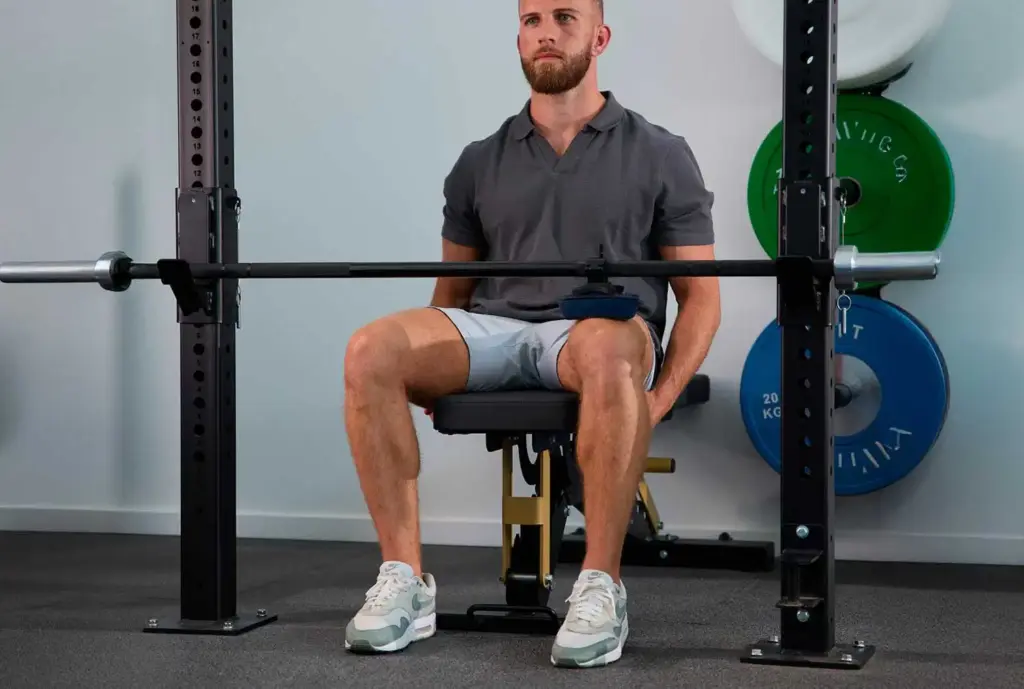 Rehabilitación específica por zona del cuerpo 43 Un atleta realizando un ejercicio de gemelos sentado en un banco de pesas con una barra, utilizando un sensor de fuerza azul Kinvent sujeto al muslo para monitorizar la fuerza muscular y la progresión.