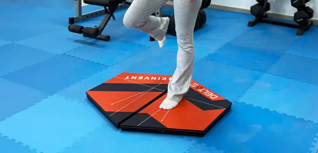 A patient performing a single-leg unilateral balance test standing on Kinvent K-Deltas XL extra-large connected force plates, inside a facility with blue gym floori