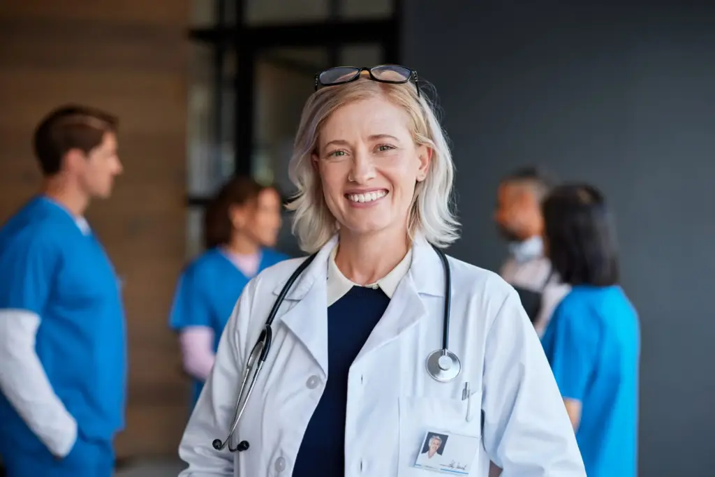 Portrait of a smiling female physician with blonde hair wearing a white lab coat and stethoscope, with her healthcare team in blue scrubs standing blurred in the background