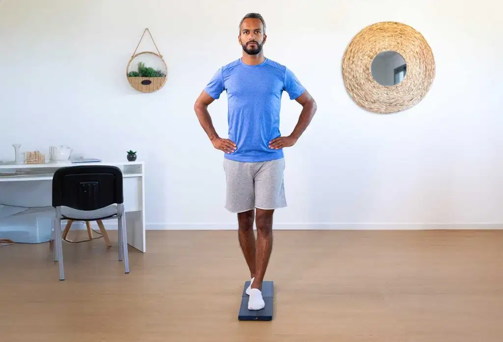 便携式测力板K-Force Plates 42 A man standing on one leg with hands on his hips, performing a single-leg balance test on a Kinvent force plate inside a bright room