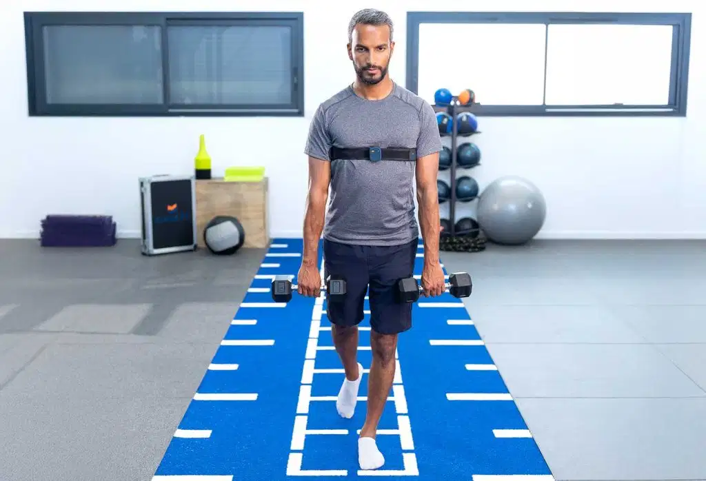 A man holding dumbbells in each hand while walking on a blue gym turf track, wearing a K-Move sensor strap across his chest