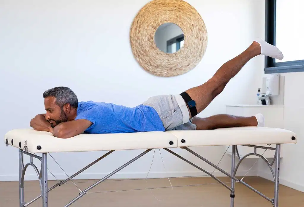 A man lying prone on a treatment table, performing a left leg raise while wearing a K-Move sensor strap on his thigh