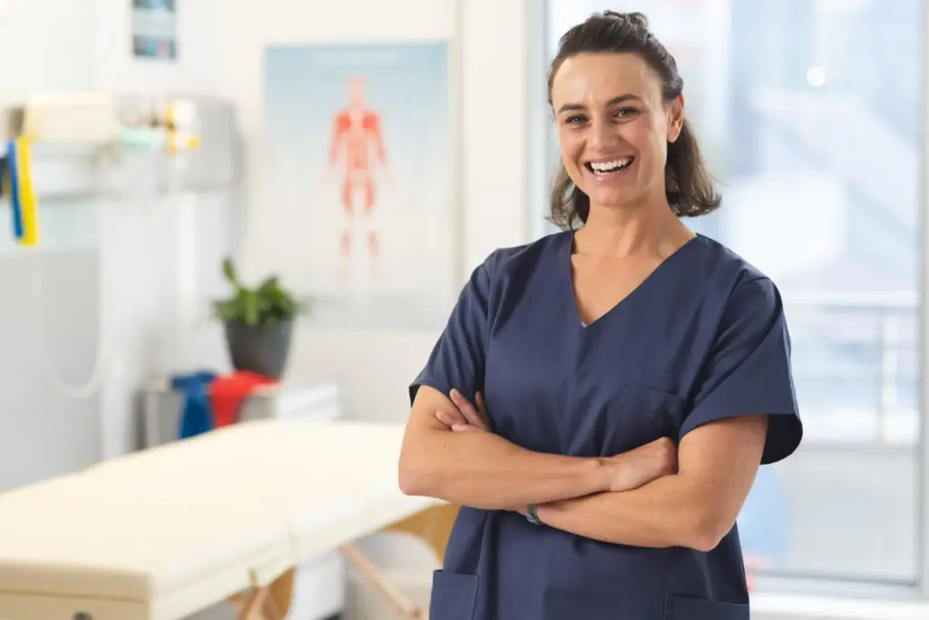 Fisioterapeuta 34 Retrato de una fisioterapeuta caucásica feliz y sonriente, de pie con los brazos cruzados y vistiendo un uniforme azul oscuro en una clínica de rehabilitación, con una camilla de tratamiento borrosa en el fondo