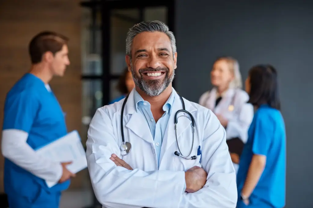 Portrait of a confident, smiling male physician with arms crossed wearing a white lab coat and stethoscope, with other healthcare team members in blue scrubs standing blurred in the background