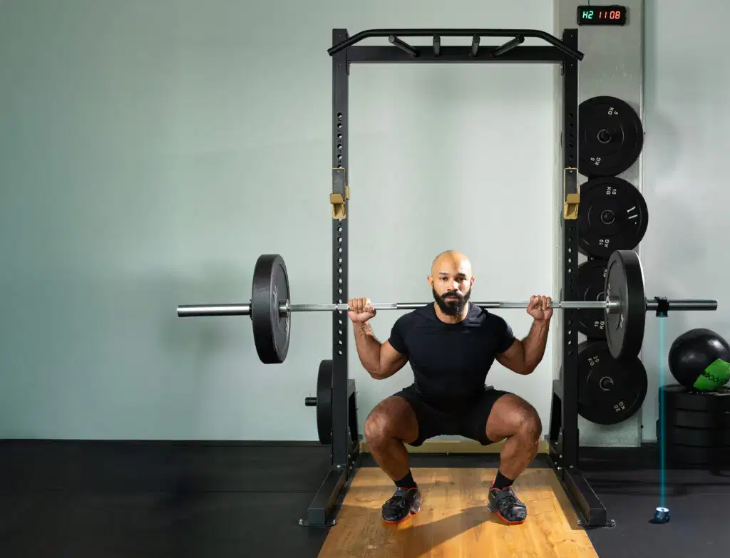 An athlete performing a barbell squat exercise in a gym cage, monitored by the Kinvent K-Power laser system placed on the floor