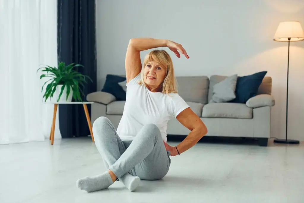 Patient at home rehabilitation 35 A senior woman sitting cross-legged on her living room floor while performing lateral trunk bending and arm stretching exercises during a home rehabilitation session